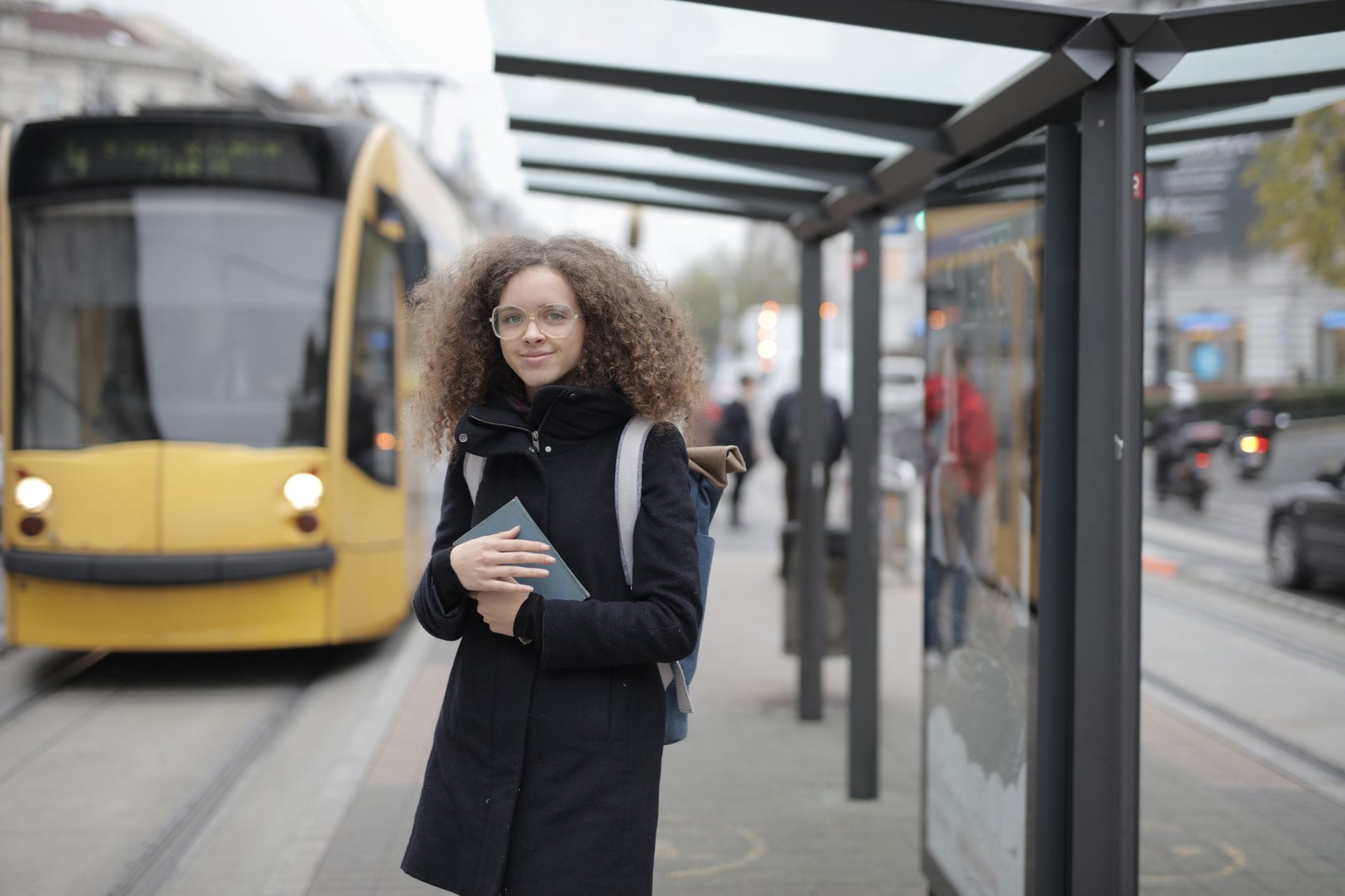 Młoda kobieta z plecakiem czeka na tramwaj z książką w ręku